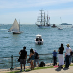 People watching ships on the water from a waterfront walkway with a large sailing ship in view.