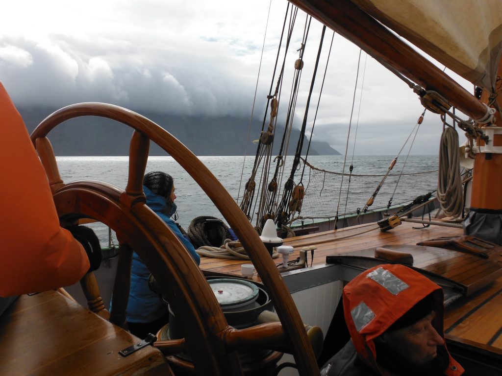 A person is sailing on a wooden ship, navigating through cloudy weather with a view of the sea and distant mountains.